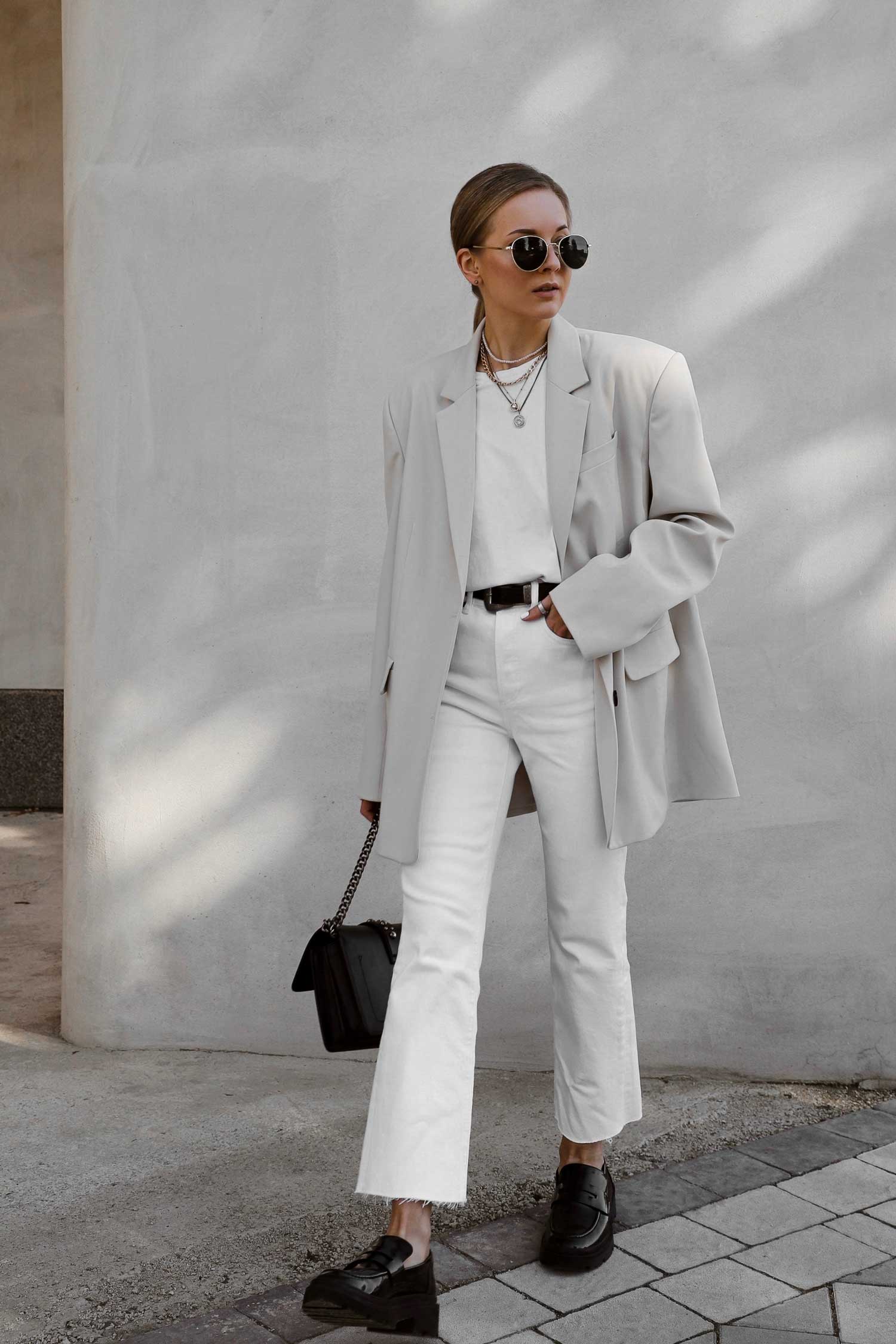 A person stands wearing a white blazer, white pants, and black shoes, accessorized with sunglasses, necklaces, and a black handbag, against a minimalist concrete wall.