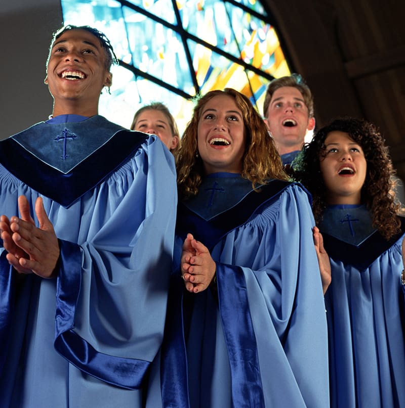 A group of choir members in blue robes sing enthusiastically in front of a stained glass window.