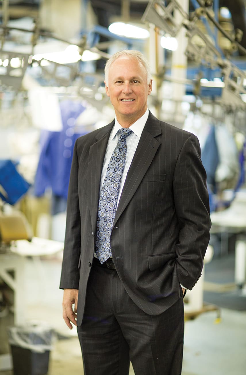 A man in a business suit stands confidently with one hand in his pocket in an industrial setting, surrounded by machinery and equipment.