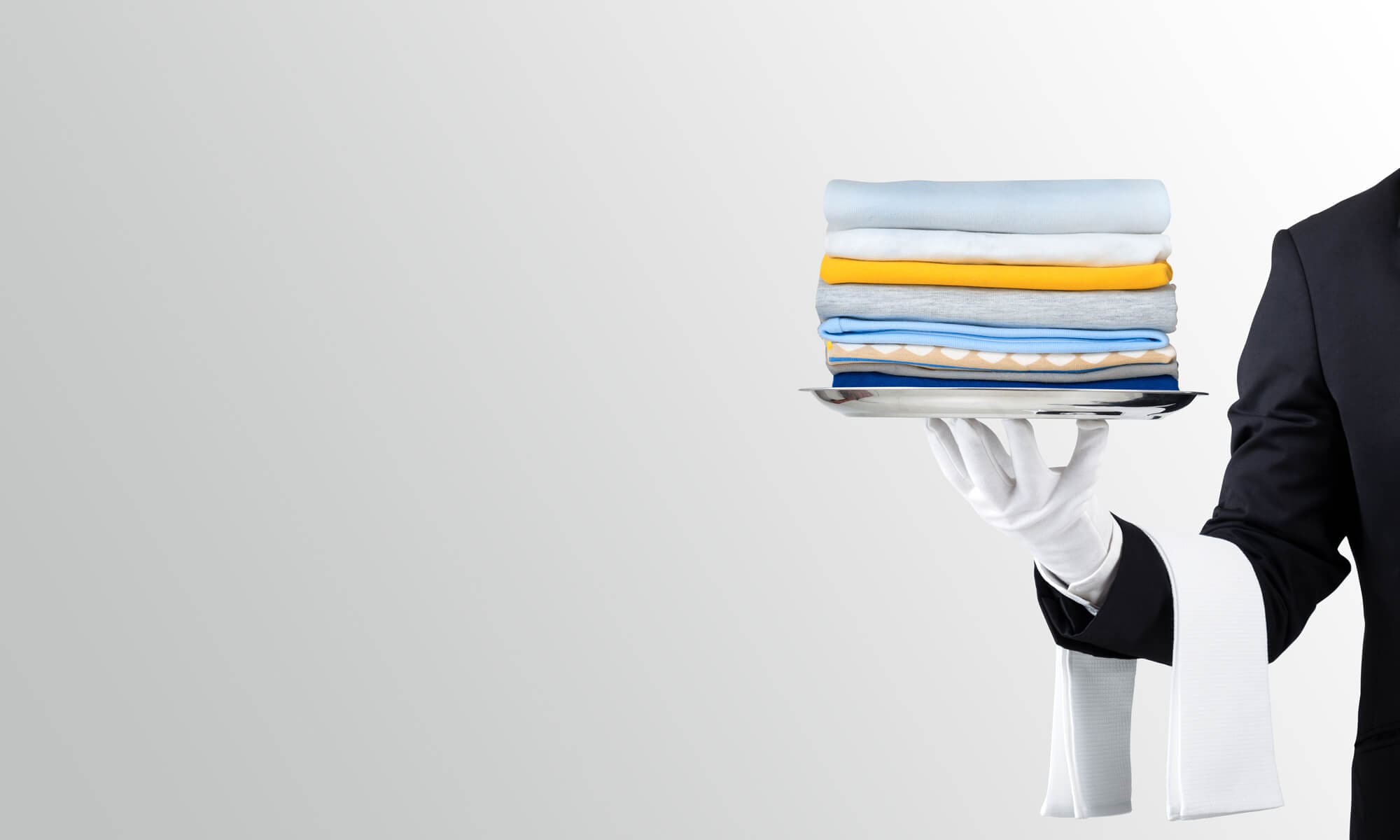A gloved hand carrying a stack of folded clothes on a silver tray against a plain background.