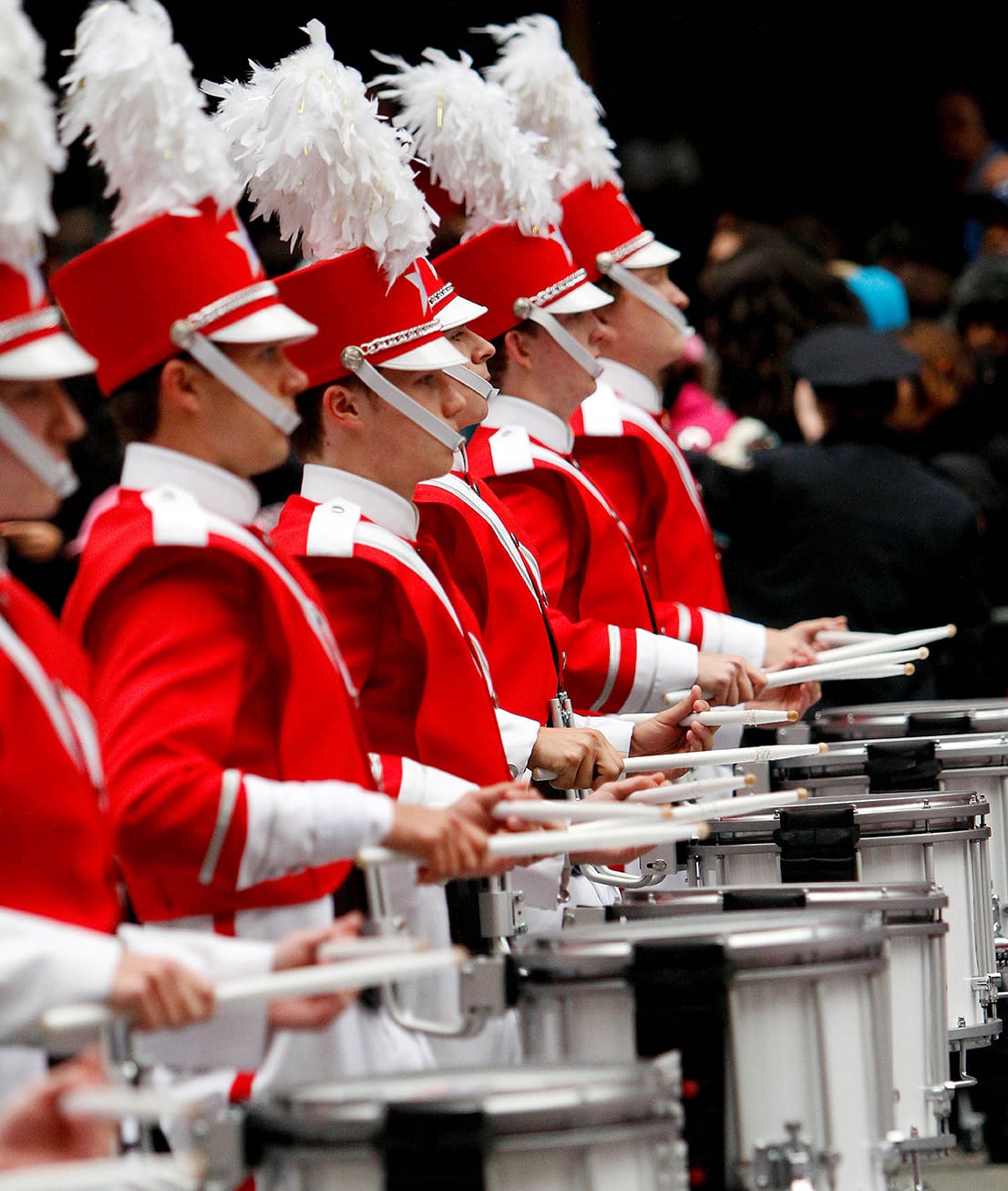 A row of marching band drummers dressed in red uniforms with white feathered hats perform in unison.