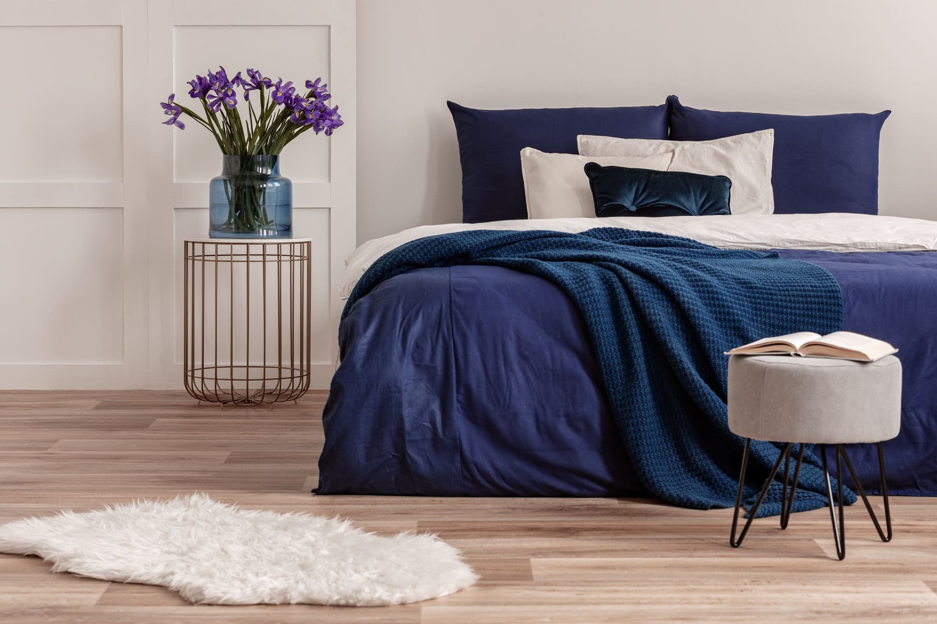 A neatly made bed with blue bedding, a small round side table holding a vase of purple flowers, and an ottoman with an open book on it. A white fluffy rug is on the wooden floor.