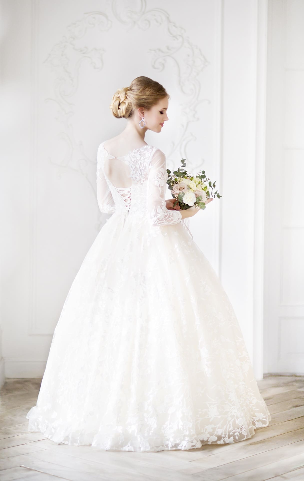 A bride in a long, white lace wedding gown with sleeves holds a bouquet and looks to the side in a softly lit room with ornate white decor.