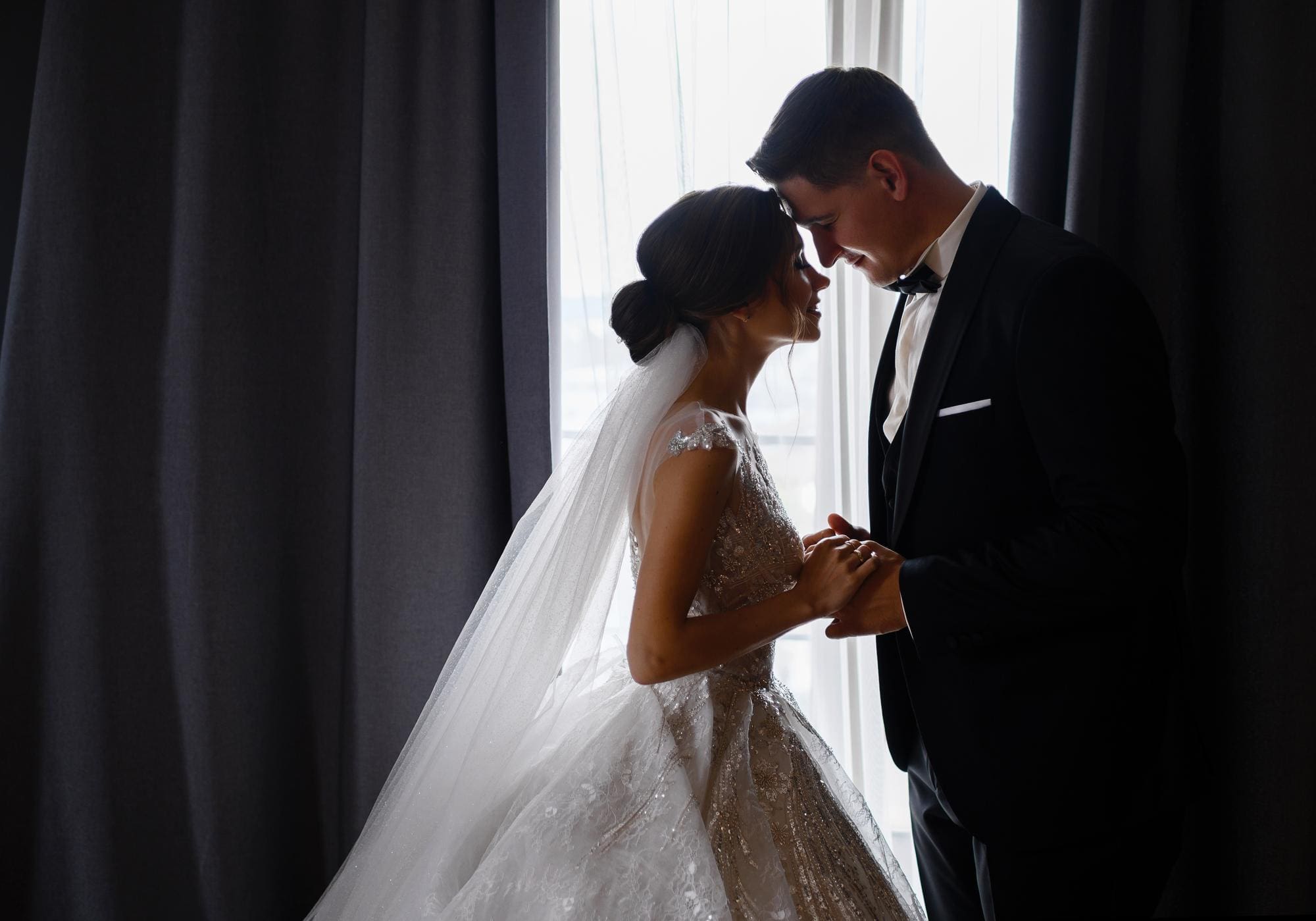 Bride and groom hold hands and lean foreheads together in front of a window, both wearing formal wedding attire.