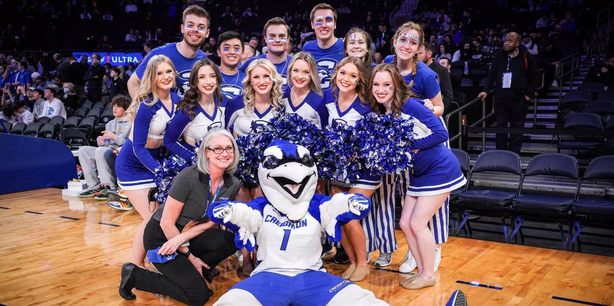 Cheerleaders and mascot pose together on a basketball court, smiling. They wear blue and white uniforms, with pom-poms held by some in the front row.