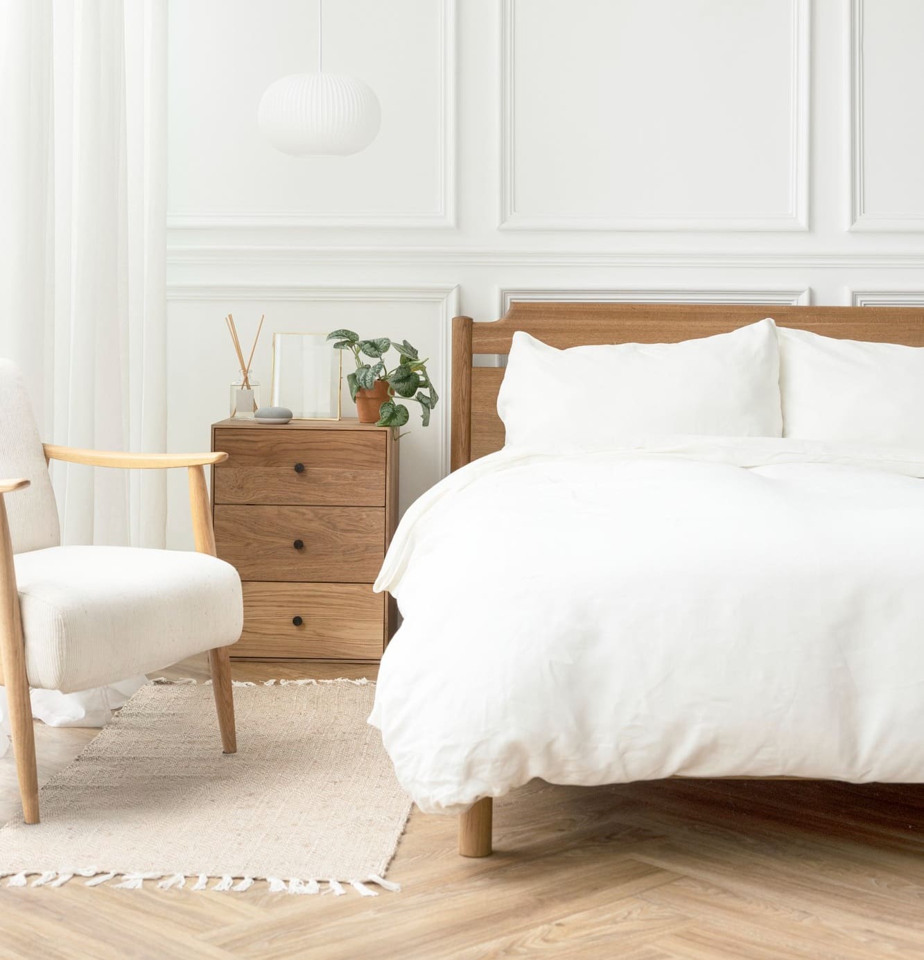 Minimalist bedroom with a wooden bed, white bedding, a wooden nightstand, and a white armchair on a textured rug. A plant, diffuser, and hanging lamp accentuate the decor.