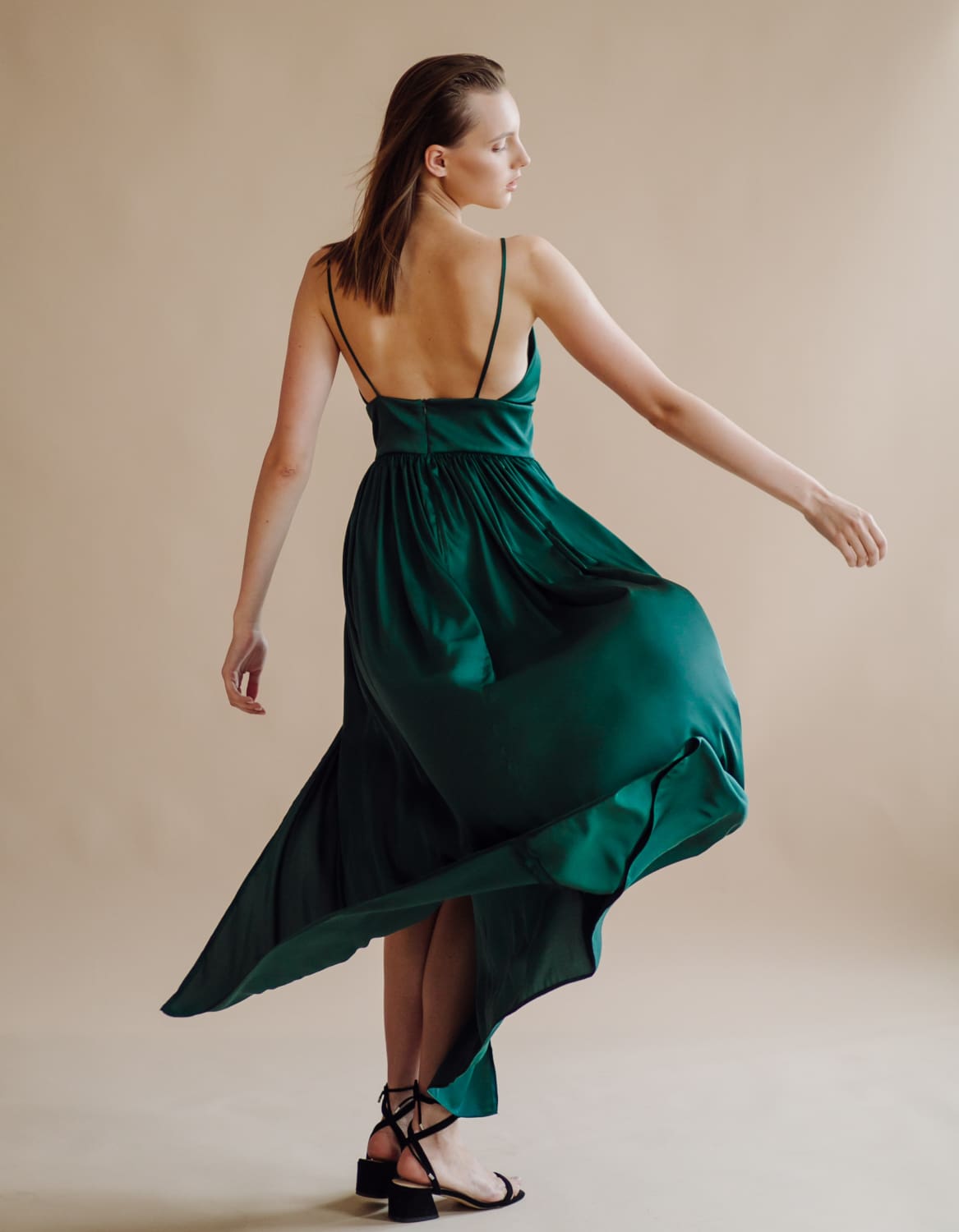 Woman in a flowing green dress with spaghetti straps, viewed from the back, standing against a neutral background.