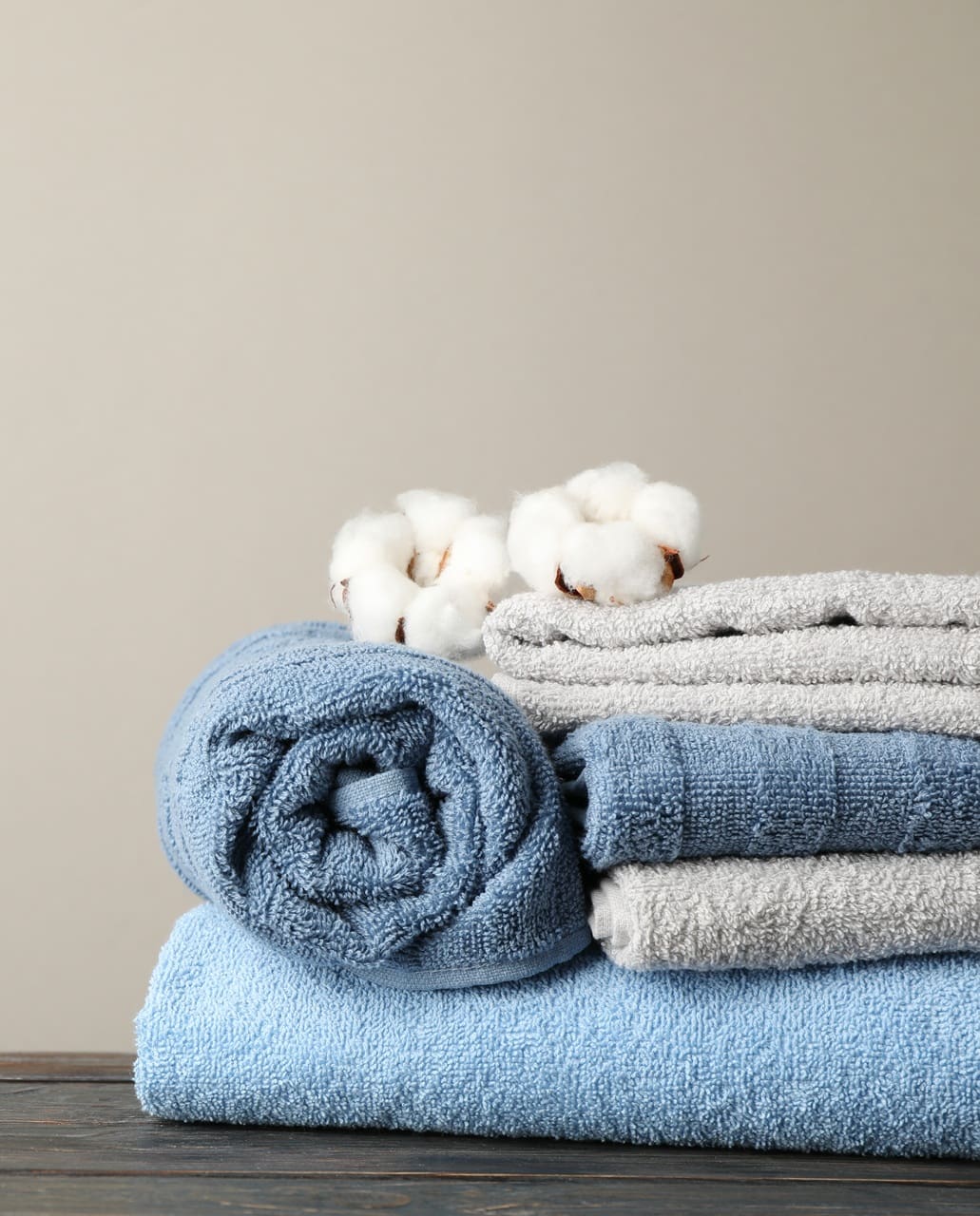 Rolled blue and folded gray towels on a table, with cotton stems placed on top. Neutral background.