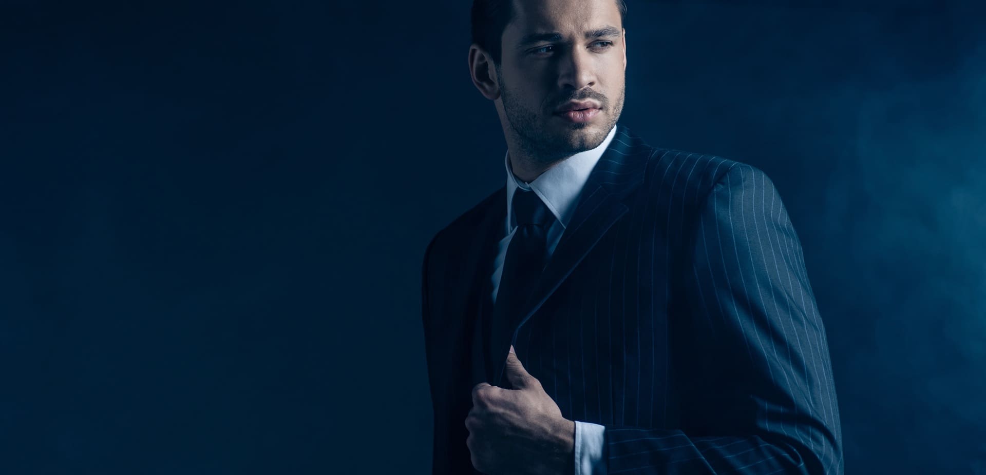 A man in a dark pinstripe suit and tie stands against a moody blue background, holding the lapel of his jacket with one hand.