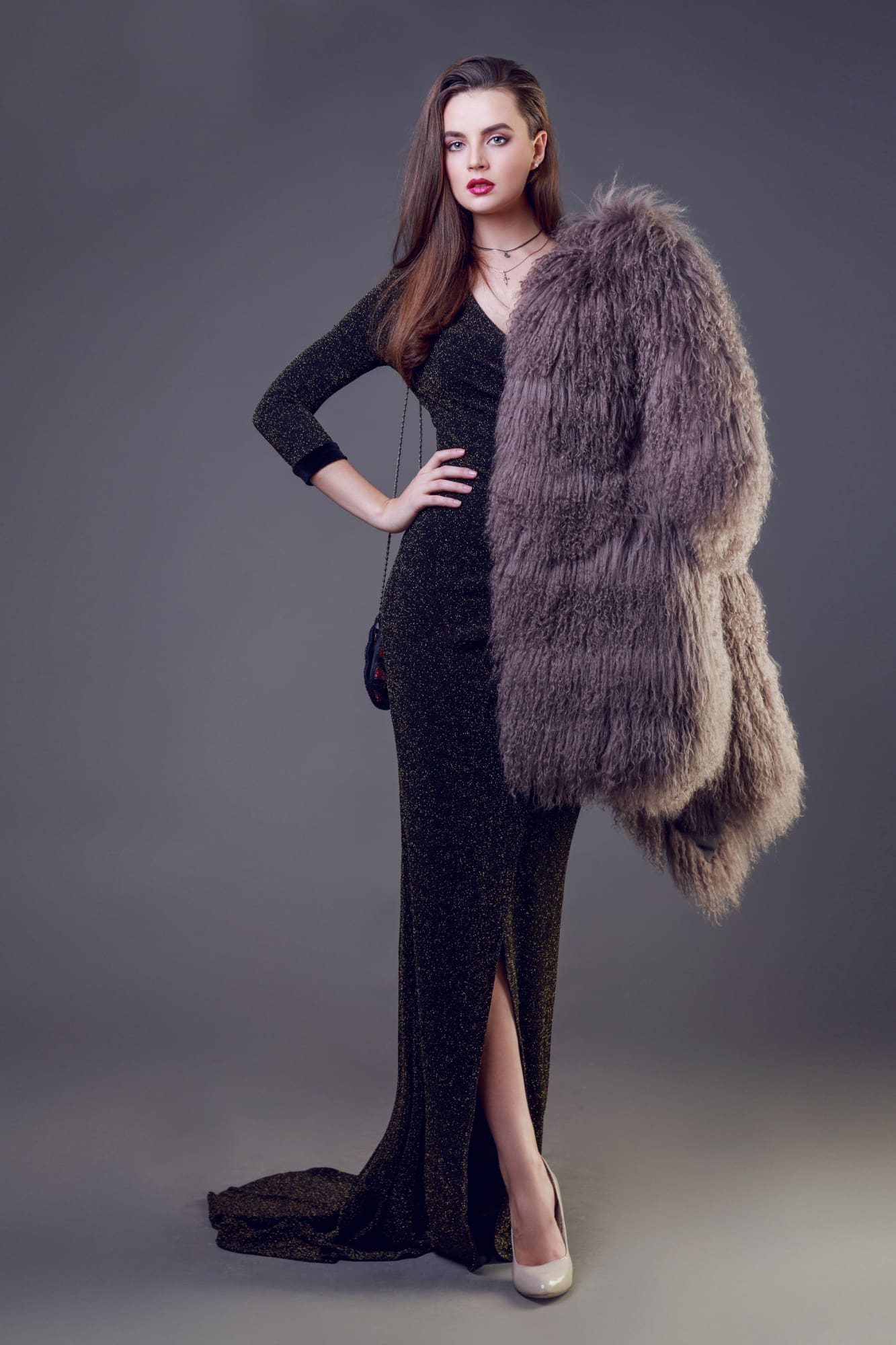 Person in a long black dress and high heels, holding a large, furry coat over one arm, stands against a plain gray background.