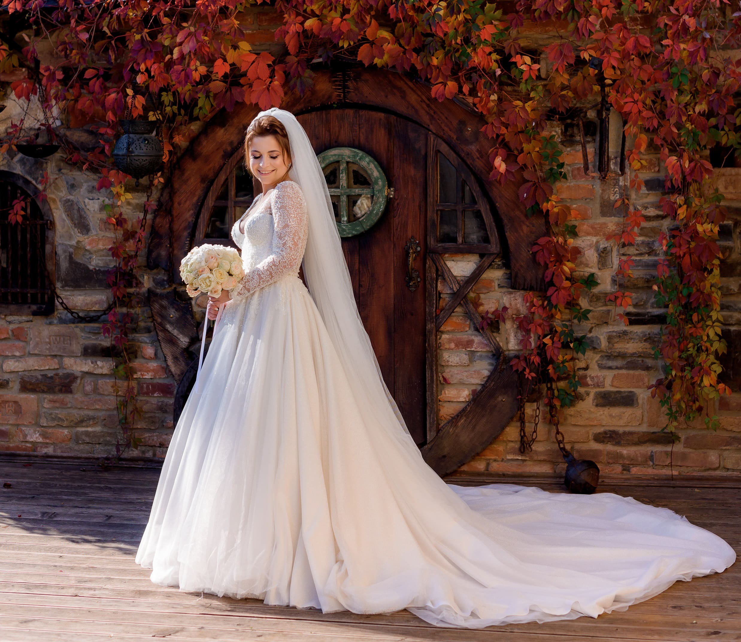 Bride in a white gown and veil stands holding a bouquet. She poses in front of a rustic wooden door, surrounded by brick walls covered with autumn leaves.