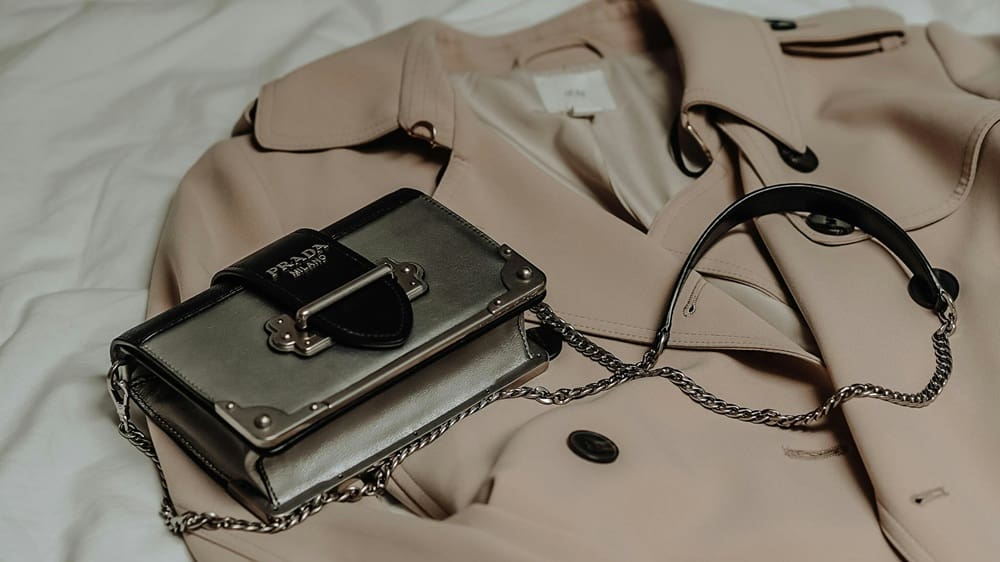 Fashion Cleaners A beige trench coat and a small black Prada handbag with a chain strap are laid out on a white surface.