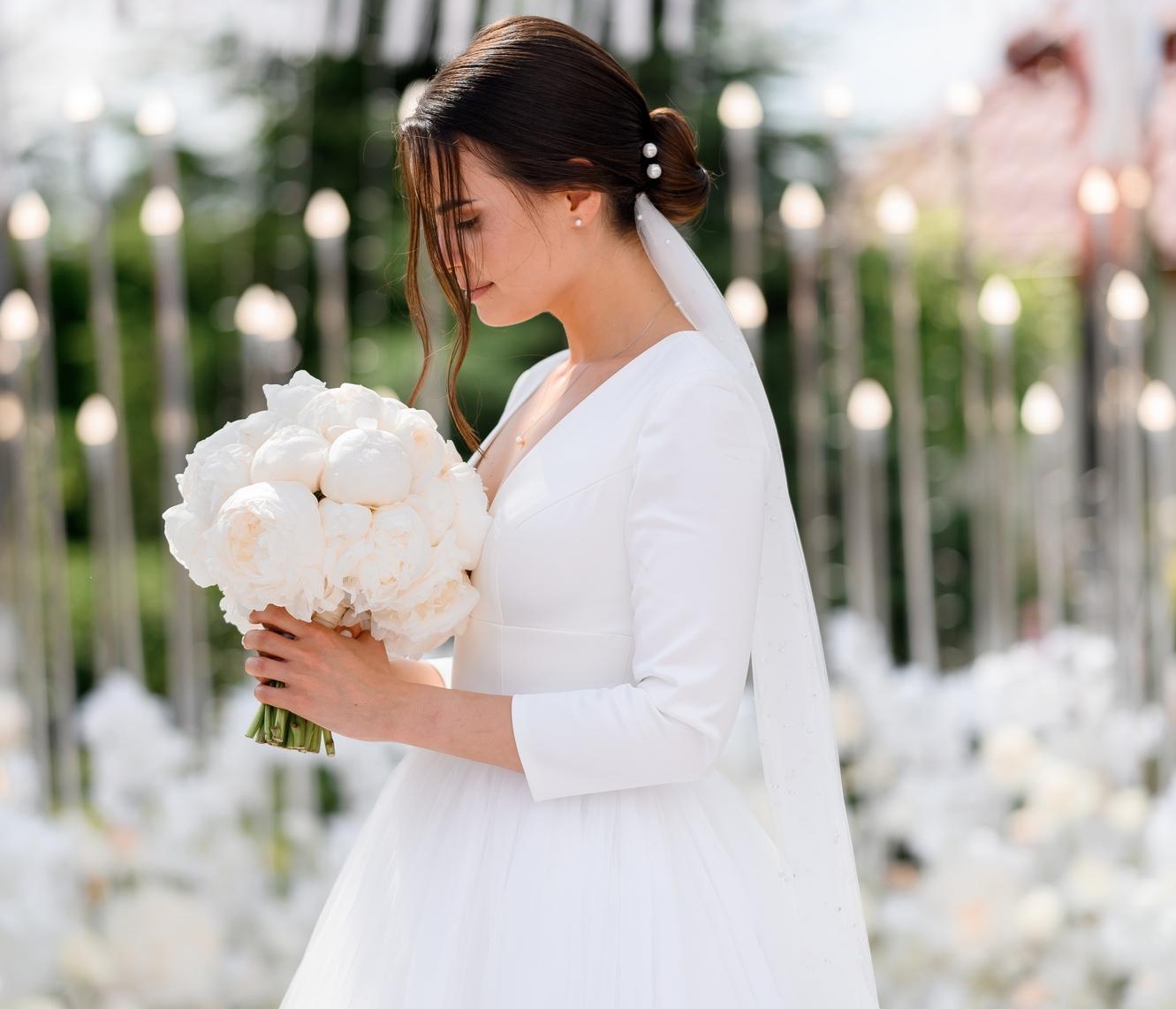Fashion Cleaners A bride in a white dress holds a bouquet of white flowers, standing outdoors with a blurred background of lights and greenery.