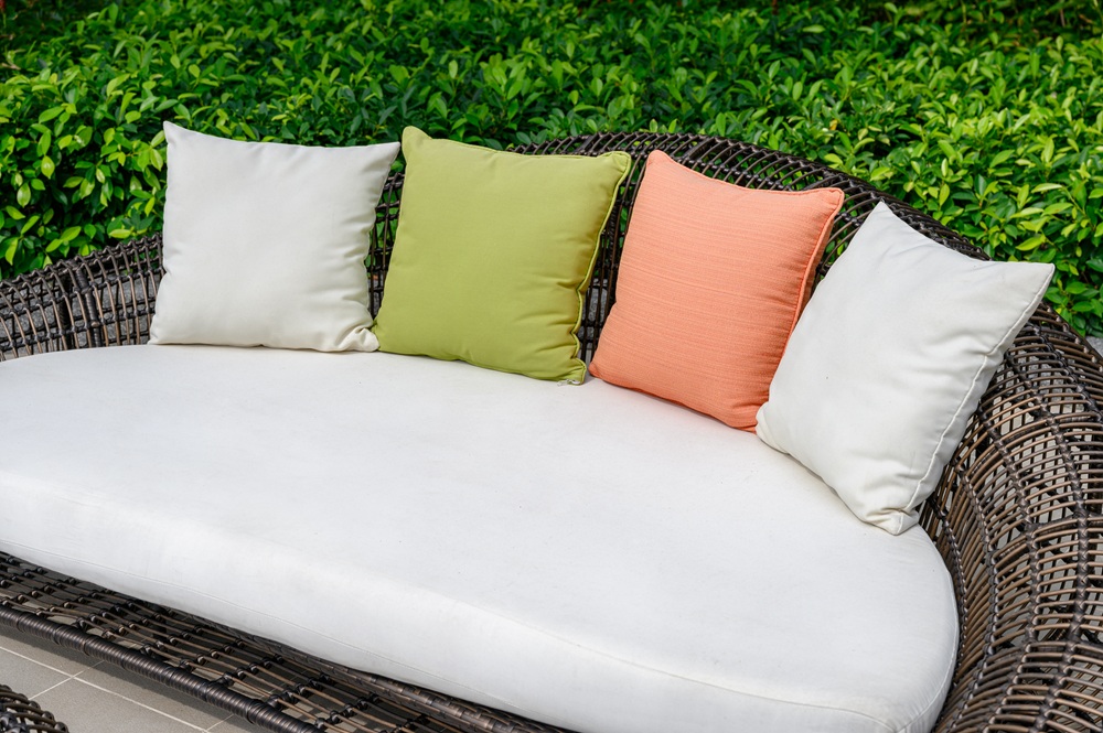 Fashion Cleaners A wicker outdoor sofa with a white cushion and four pillows—two white, one green, and one orange—set in front of green shrubs.
