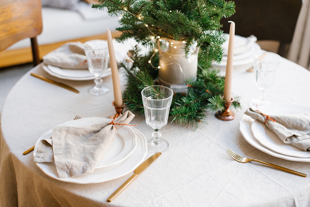 Fashion Cleaners A round table set for three with white plates, linen napkins, gold utensils, glasses, two candles, and a small pine tree centerpiece with lights.