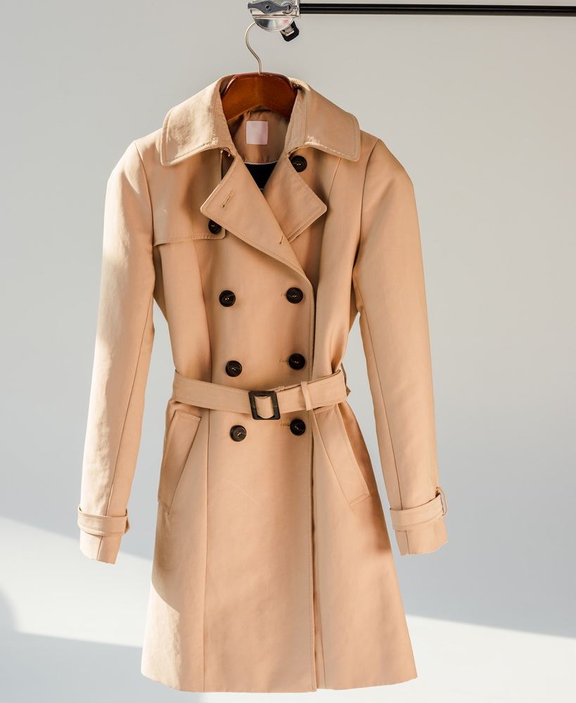 Fashion Cleaners A beige double-breasted trench coat with a belt hangs on a wooden hanger against a light background.